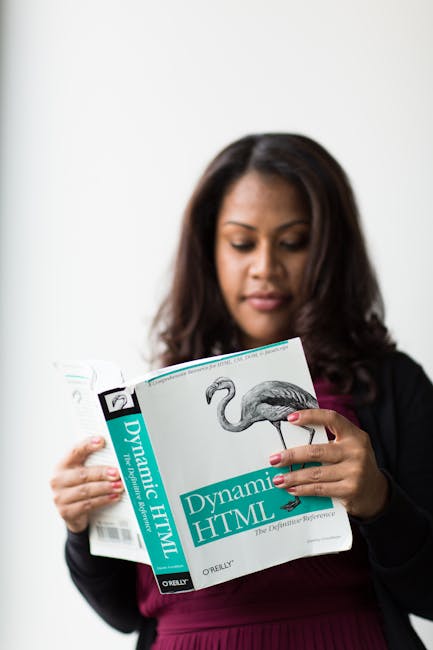 A woman with dark hair and medium skin tone is holding and reading a book titled 'Dynamic HTML' in a well-lit room with a plain white wall background. She is dressed in a dark blazer over a purple top