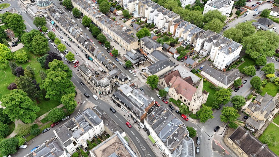 An aerial view of Barnes High Street in a residential area, showing a mix of shops, cafes, and houses. The street features multiple lanes with parked cars along both sides, and finished surfaces inclu