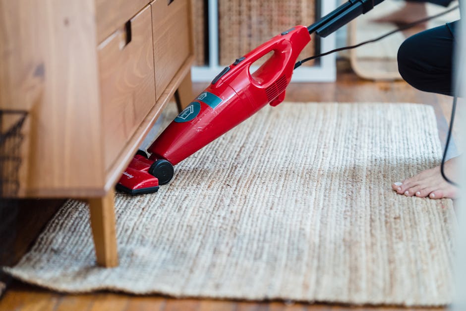 Close-up image of a person vacuuming an ornate, patterned carpet in a well-lit room. The vacuum cleaner has a black head and a long black hose, with the owner wearing dark pants and white footwear. Th